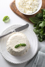 Fresh ricotta (cream cheese) with basil and knife on white wooden table, flat lay