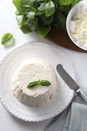 Photo of Fresh ricotta (cream cheese) with basil and knife on white wooden table, flat lay