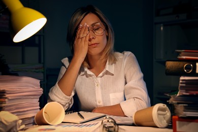 Overwhelmed woman surrounded by documents and paper coffee cups at table in office at night