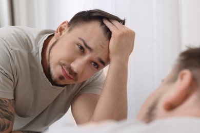 Baldness concept. Sad man with receding hairline looking at mirror indoors