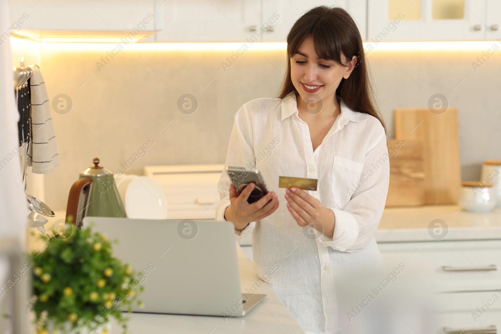 Photo of Online banking. Smiling woman with credit card and smartphone paying purchase in kitchen