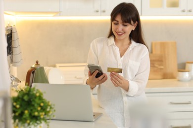 Online banking. Smiling woman with credit card and smartphone paying purchase in kitchen