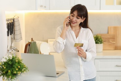 Online banking. Smiling woman with credit card talking by smartphone in kitchen