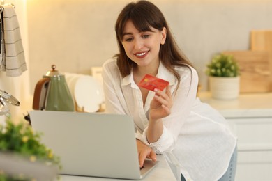 Online banking. Smiling woman with credit card and laptop paying purchase in kitchen