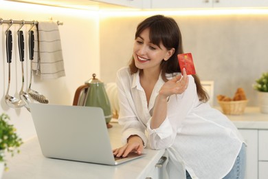 Photo of Online banking. Smiling woman with credit card and laptop paying purchase in kitchen