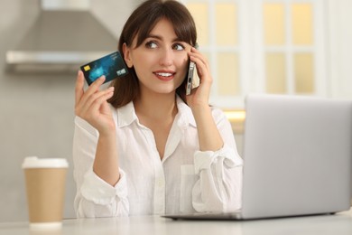 Online banking. Smiling woman with credit card talking by smartphone at table indoors