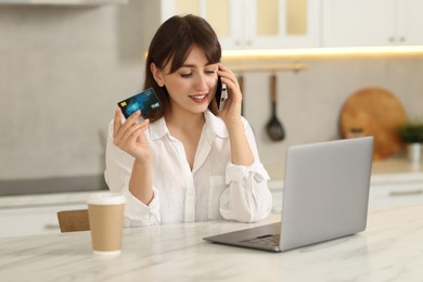 Online banking. Smiling woman with credit card talking by smartphone at table indoors