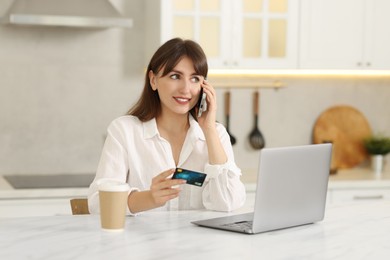 Online banking. Smiling woman with credit card talking by smartphone at table indoors