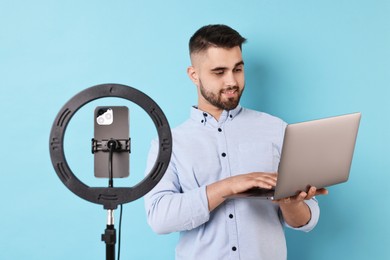 Technology blogger reviewing laptop and recording video with smartphone and ring lamp on light blue background