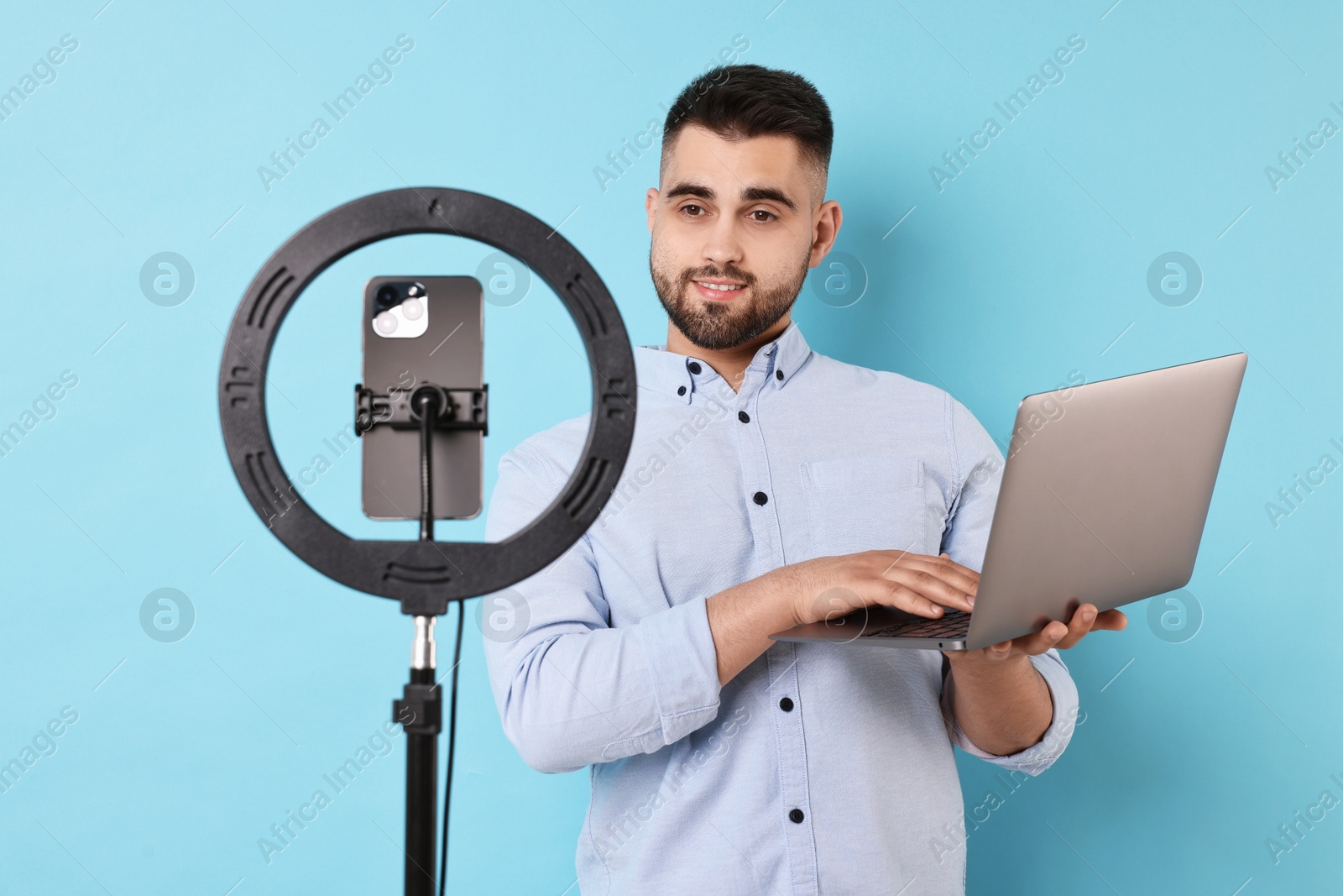 Photo of Technology blogger reviewing laptop and recording video with smartphone and ring lamp on light blue background