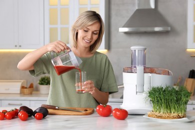 Photo of Smiling woman pouring tomato juice into glass in kitchen. Juicer and fresh products on white marble table