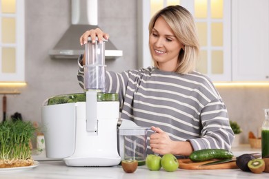 Smiling woman with fresh products using juicer at white marble table in kitchen