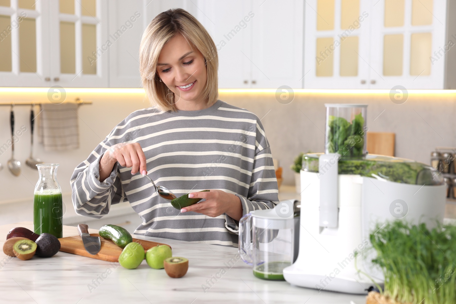 Photo of Smiling woman with fresh products using juicer at white marble table in kitchen