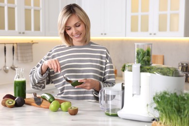 Smiling woman with fresh products using juicer at white marble table in kitchen