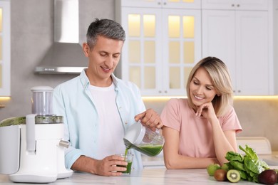 Happy couple with juicer, fresh products and tasty juice at white marble table in kitchen