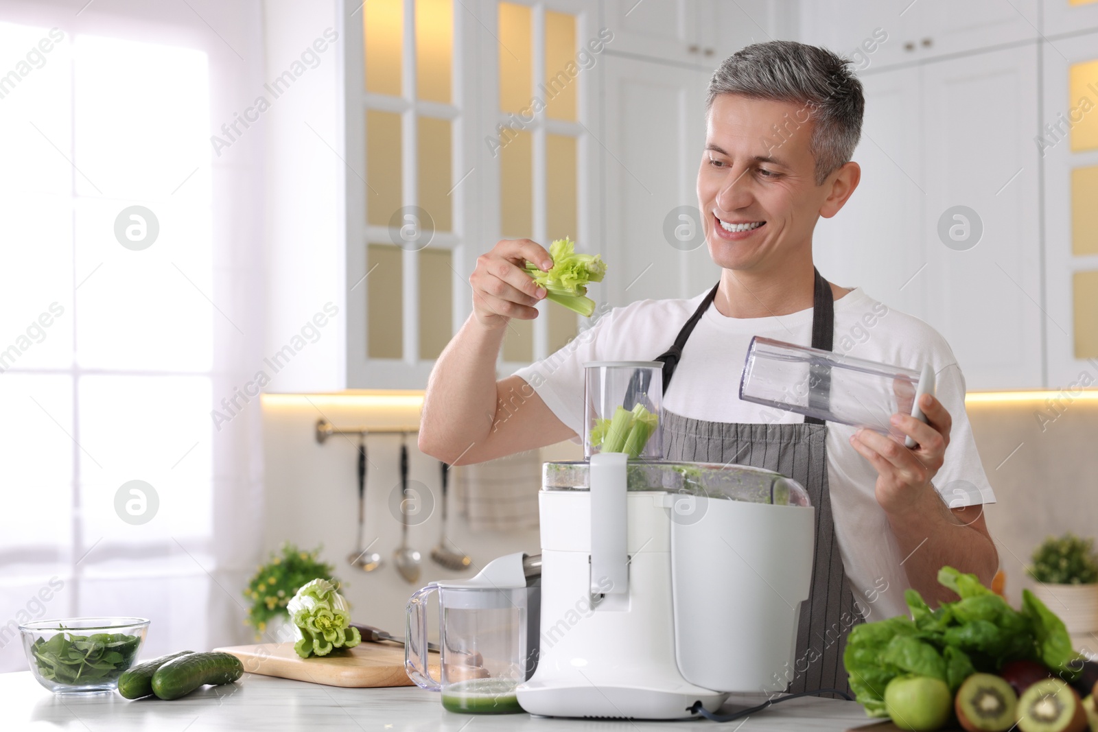 Photo of Smiling man putting fresh celery into juicer at white marble table in kitchen