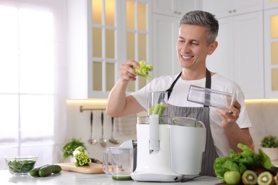 Smiling man putting fresh celery into juicer at white marble table in kitchen