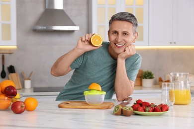 Photo of Making juice. Smiling man with orange at white marble table in kitchen
