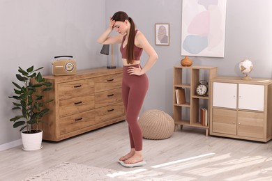 Upset woman standing on floor scale at home