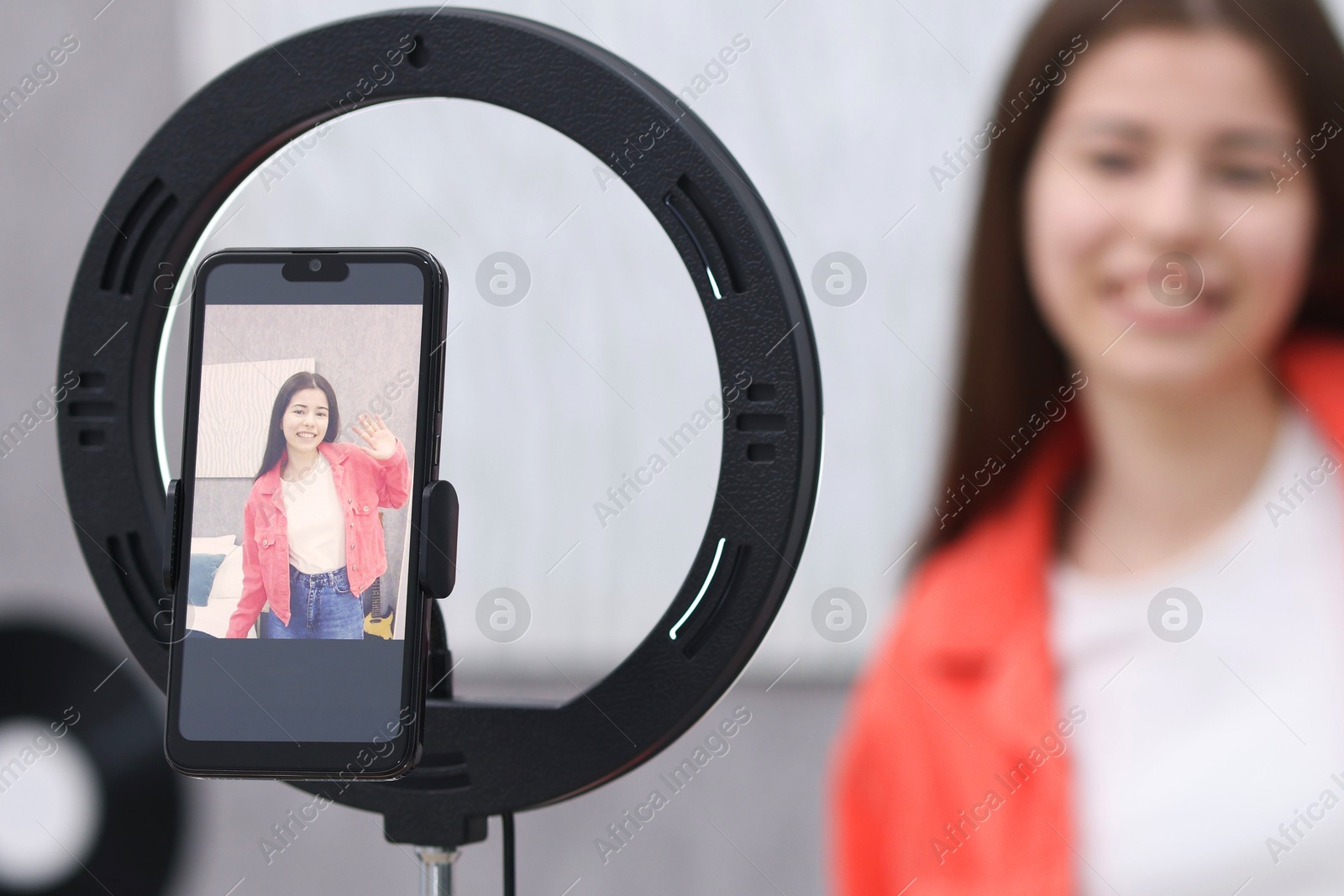 Photo of Smiling teenage blogger waving hello to her subscribers while streaming at home, focus on smartphone