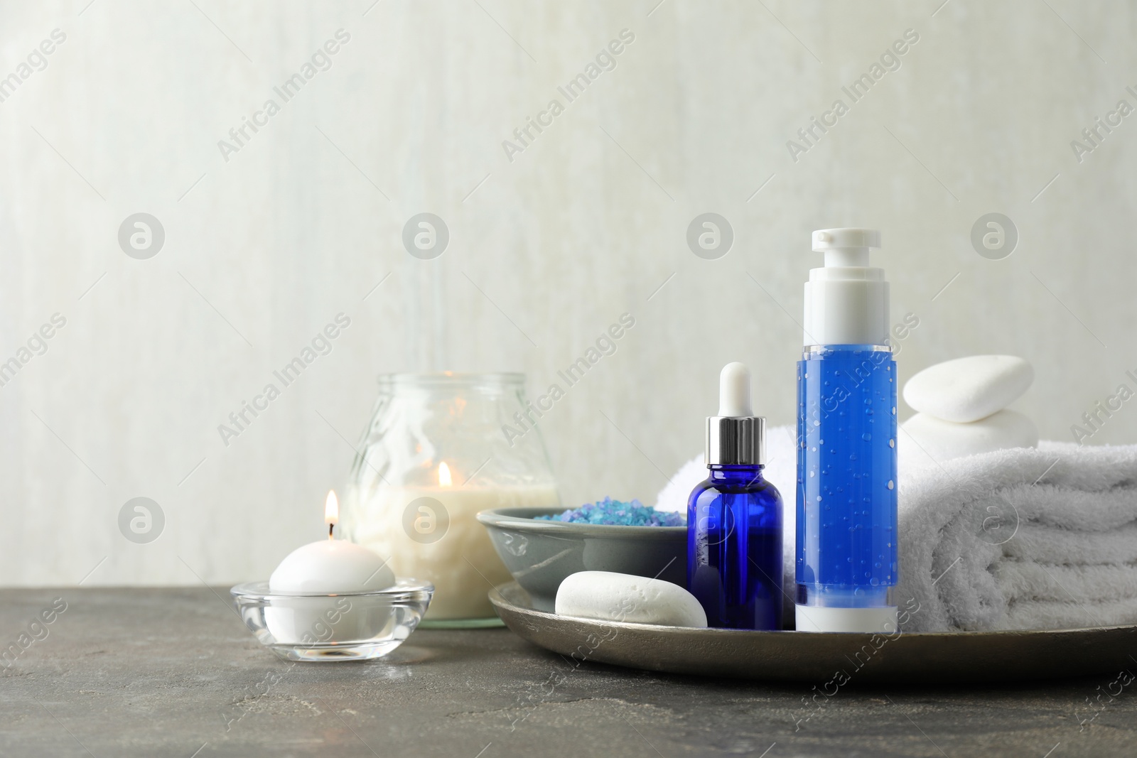 Photo of Spa composition. Cosmetic products, terry towel, burning candles and sea salt on gray table against light background, space for text