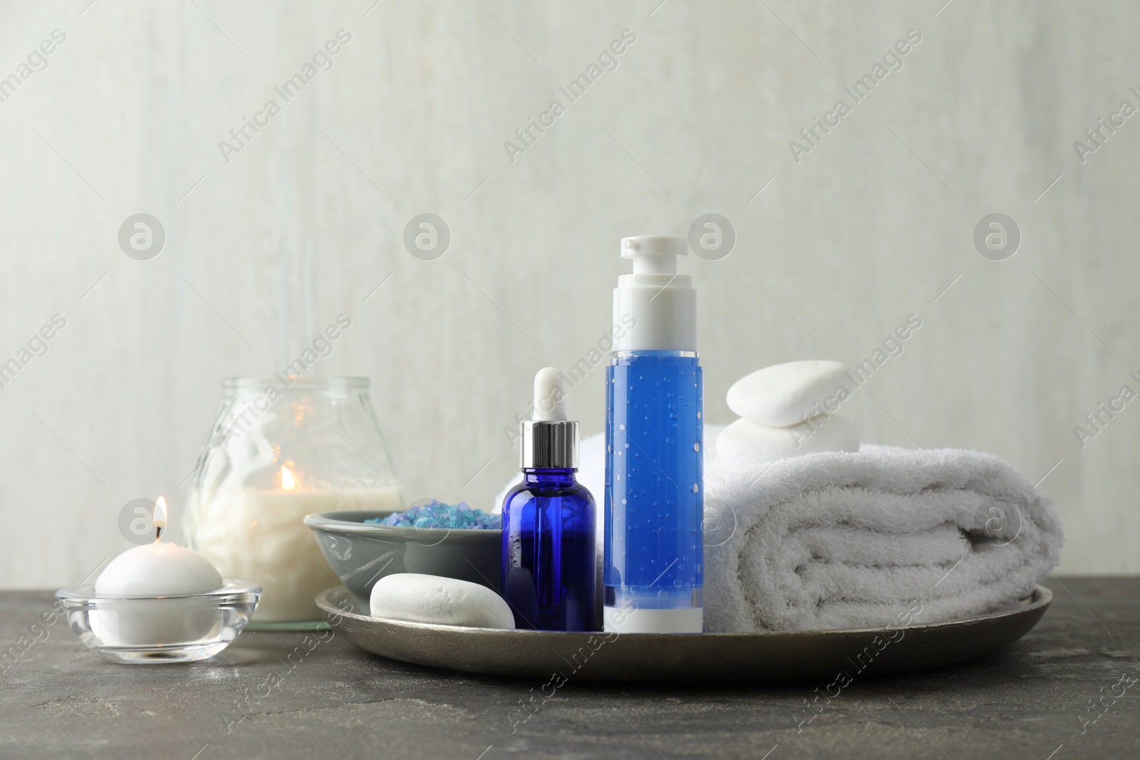 Photo of Spa composition. Cosmetic products, terry towel, burning candles and sea salt on gray table against light background