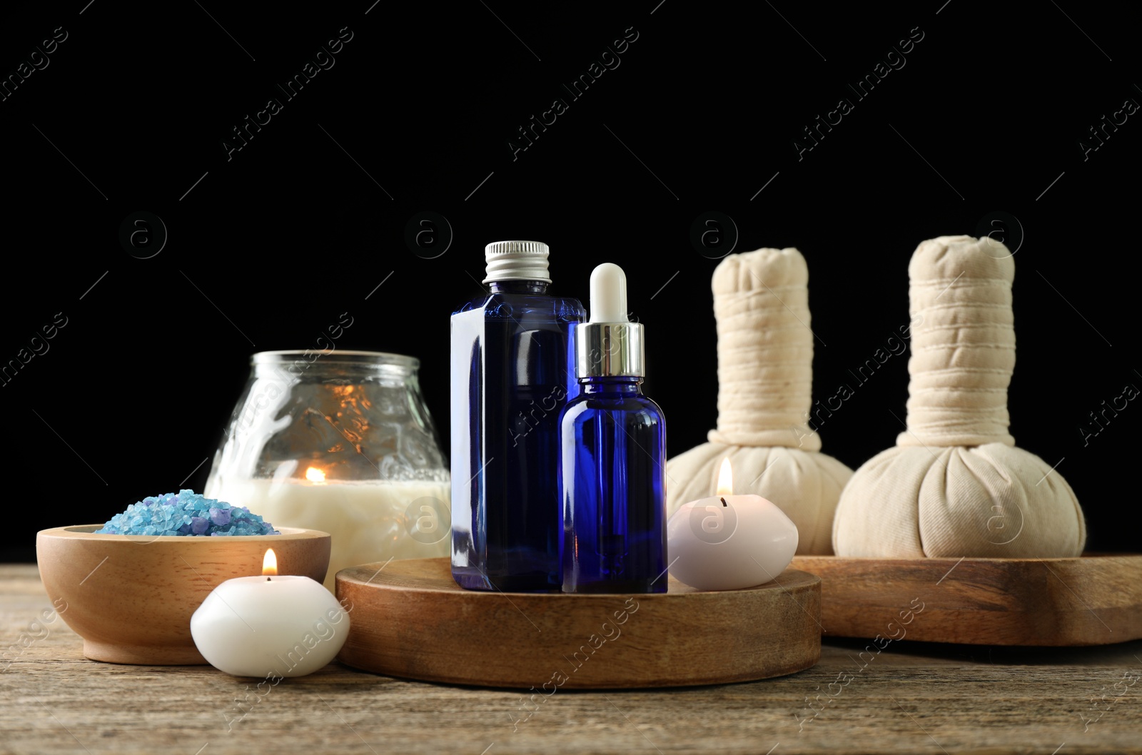 Photo of Spa composition. Cosmetic products, herbal bags, burning candles and sea salt on wooden table against black background