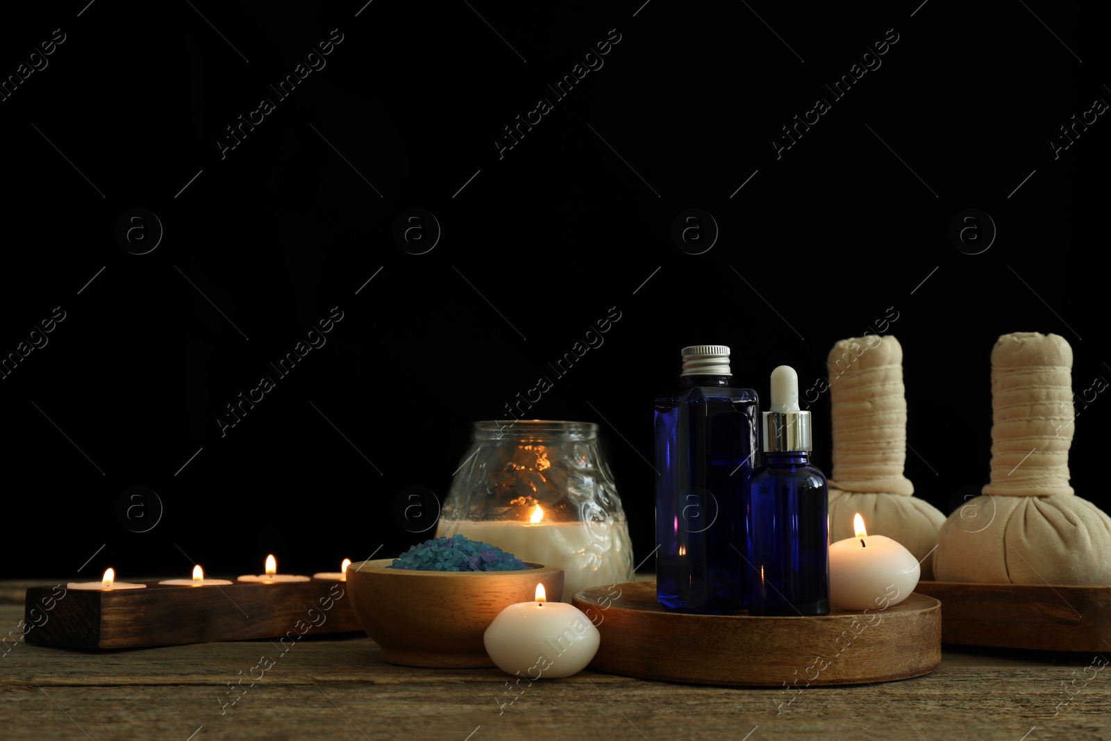 Photo of Spa composition. Cosmetic products, herbal bags, burning candles and sea salt on wooden table against black background, space for text