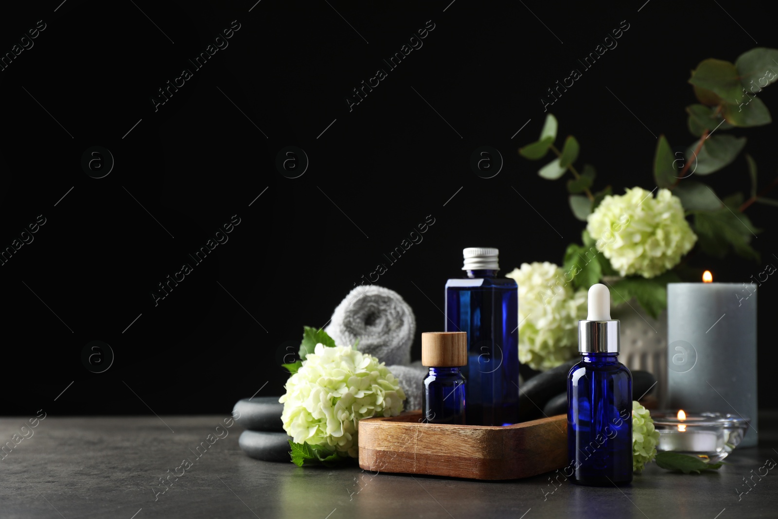 Photo of Spa composition. Cosmetic products, burning candles and hydrangea flowers on gray table against black background, space for text