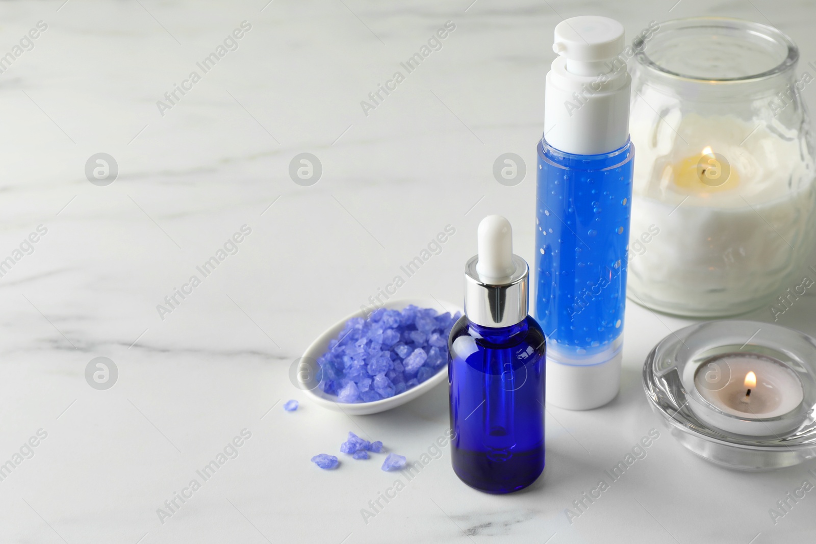 Photo of Spa composition. Bottles of cosmetic products, sea salt and burning candles on white marble table, space for text