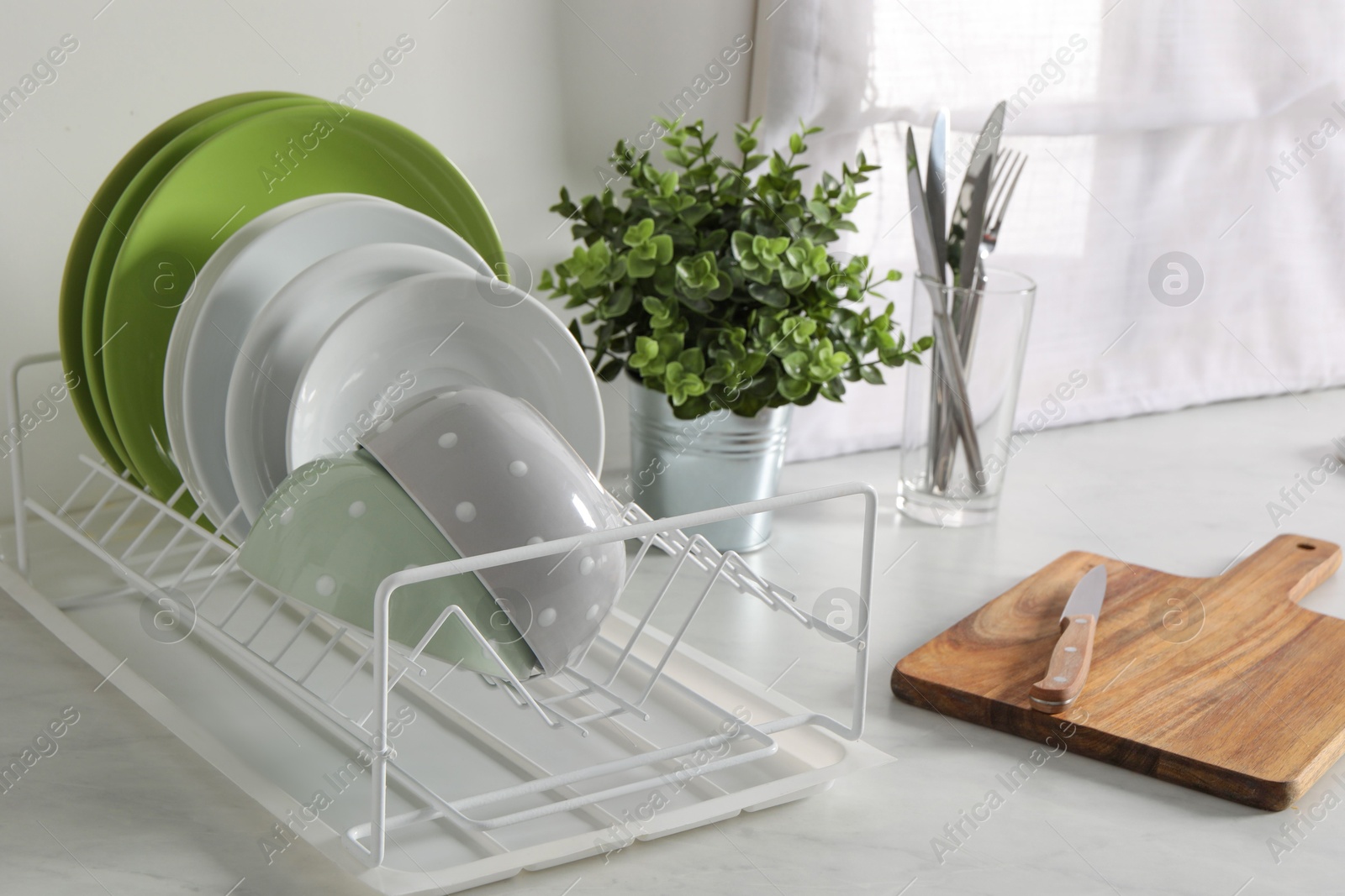 Photo of Drainer with different clean dishware and houseplant on light table in kitchen