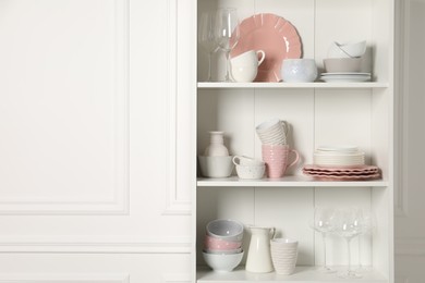 Different ceramic dishware and glasses on shelves in cabinet indoors. Space for text