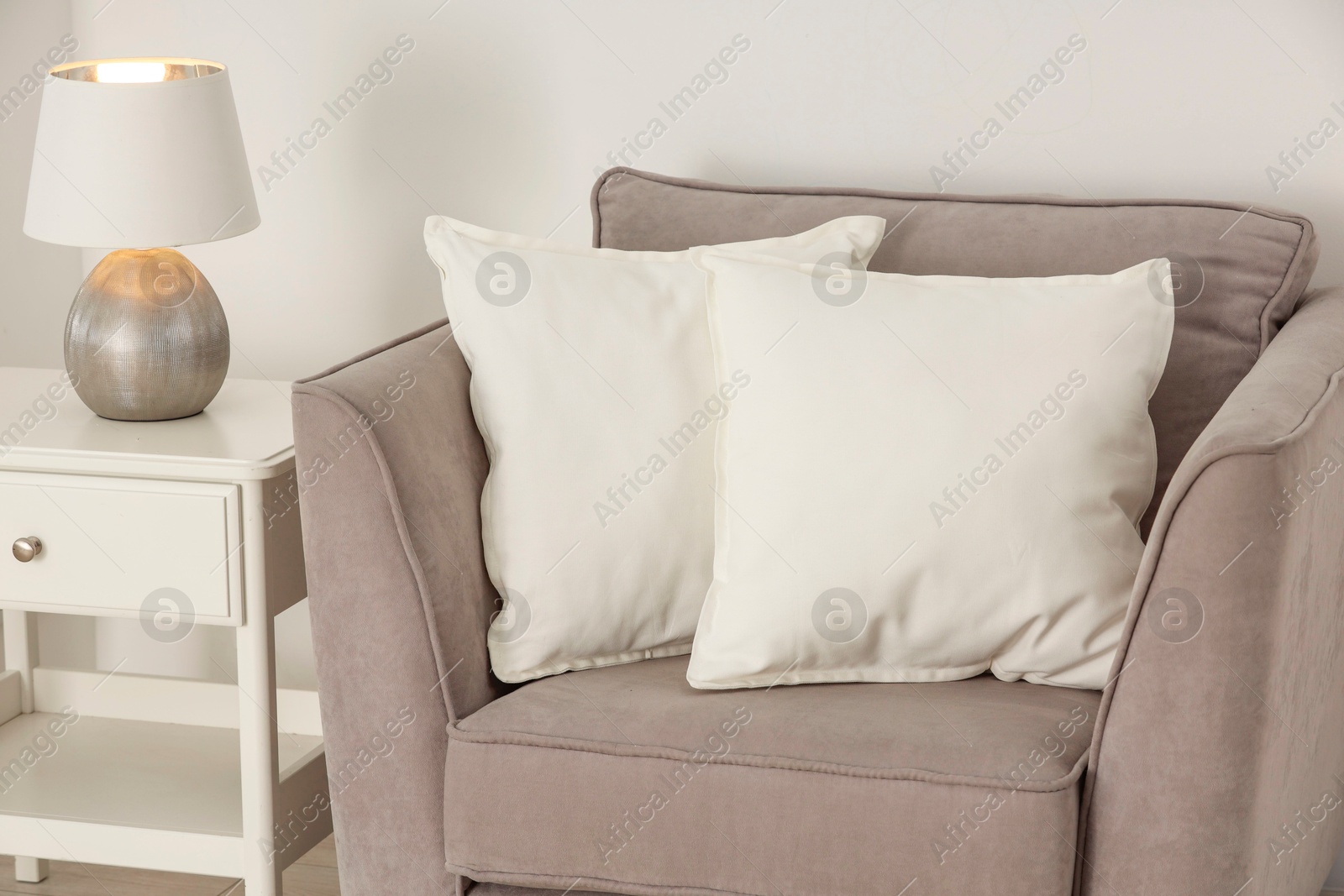 Photo of Soft white pillows on armchair, bedside table and lamp near light wall indoors