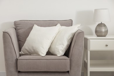 Photo of Soft white pillows on armchair, bedside table and lamp near light wall indoors