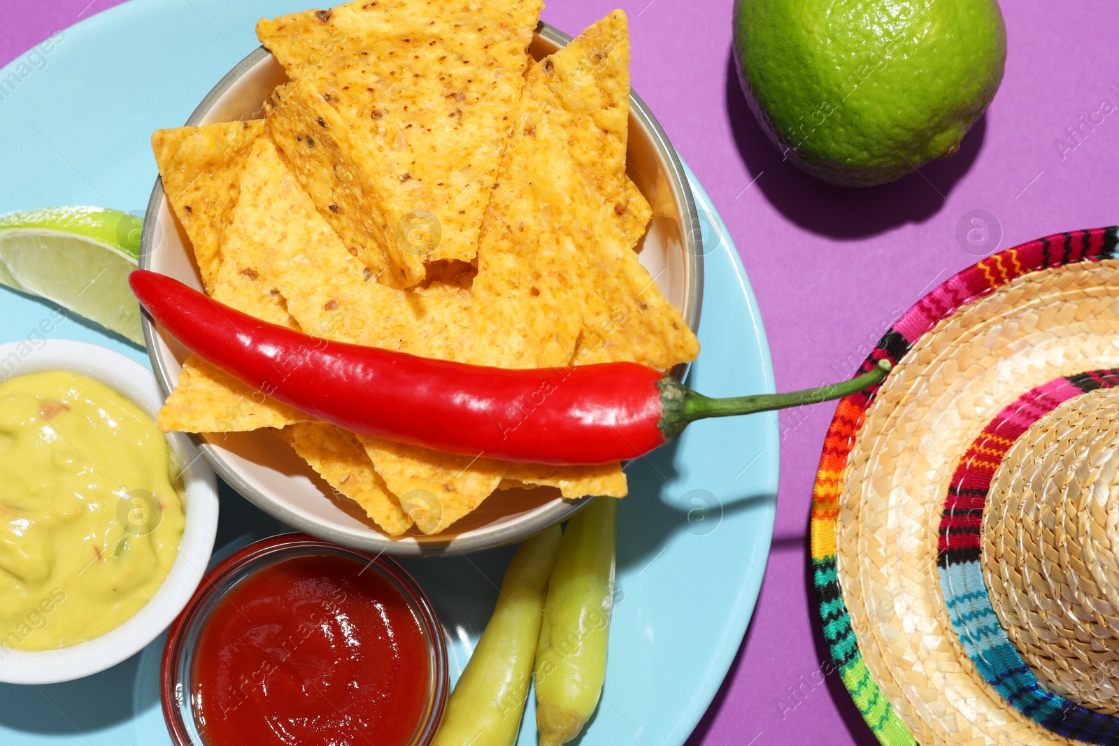 Photo of Nachos chips, guacamole, chili pepper, lime and Mexican sombrero hat on purple background, top view