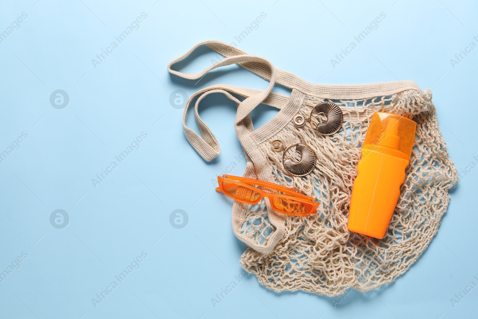 Photo of String bag, sunscreen, sunglasses and earrings on light blue background, top view. Space for text