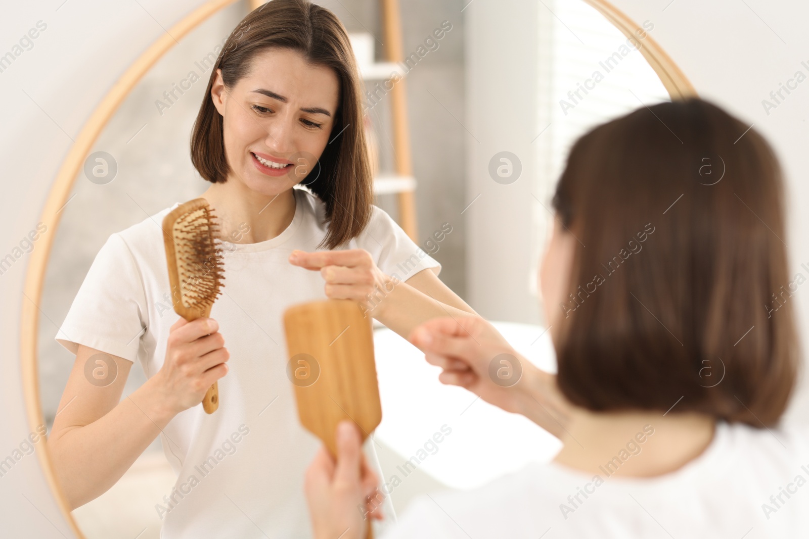 Photo of Emotional woman taking her lost hair from brush near mirror indoors. Alopecia problem