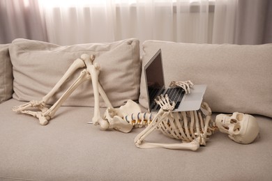 Waiting concept. Human skeleton with laptop on sofa indoors