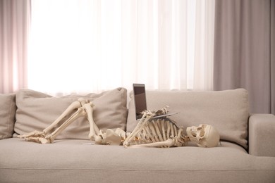 Waiting concept. Human skeleton with laptop on sofa indoors
