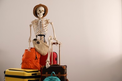 Waiting concept. Human skeleton in hat with suitcases near light wall, space for text