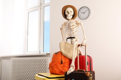 Photo of Waiting concept. Human skeleton in hat with suitcases indoors