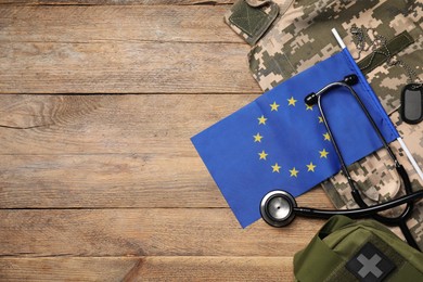 Photo of Stethoscope, flag of European Union, first aid kit and military uniform on wooden table, flat lay. Space for text