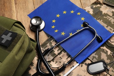 Stethoscope, flag of European Union, first aid kit and military uniform on wooden table, closeup