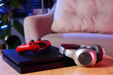 Photo of Video game console, wireless controller and headphones on wooden table indoors
