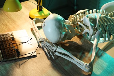 Photo of Waiting concept. Human skeleton sleeping at wooden table with laptop