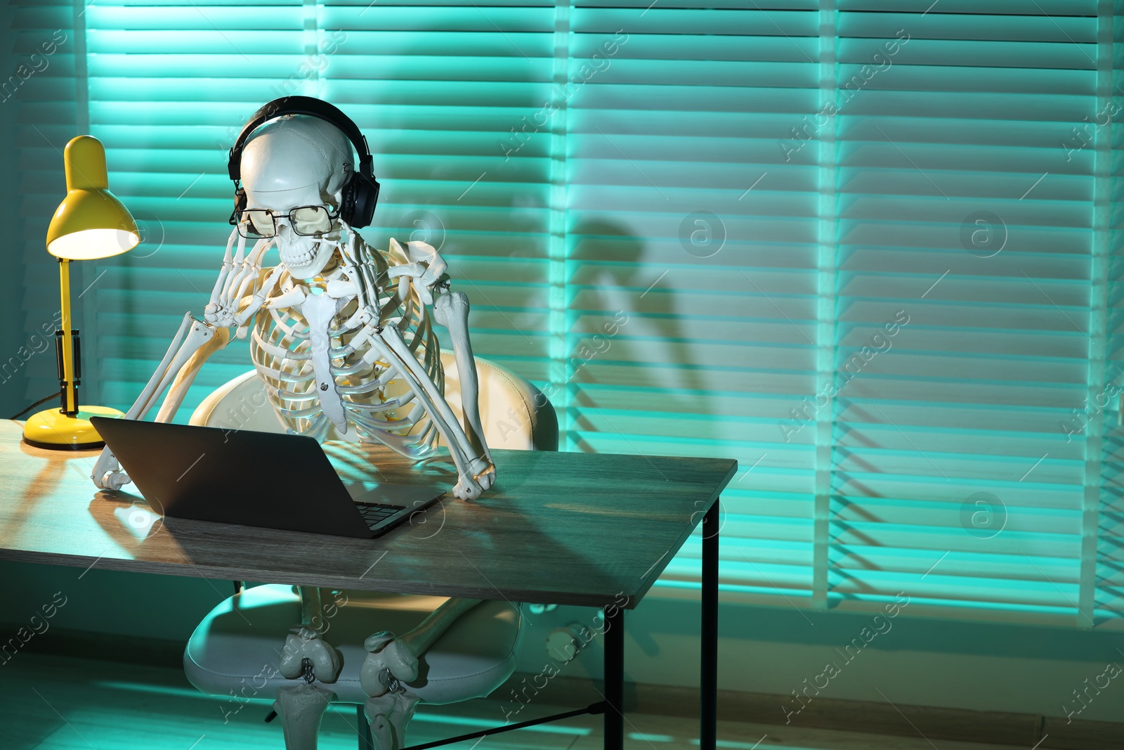 Photo of Waiting concept. Human skeleton sitting at wooden table with laptop and headphones indoors, space for text
