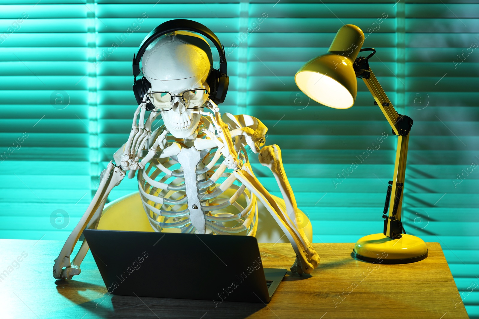 Photo of Waiting concept. Human skeleton sitting at wooden table with laptop and headphones indoors