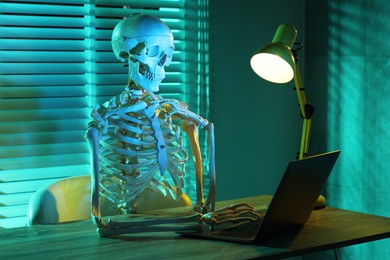 Photo of Waiting concept. Human skeleton sitting at wooden table with laptop indoors