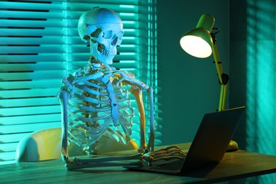 Photo of Waiting concept. Human skeleton sitting at wooden table with laptop indoors