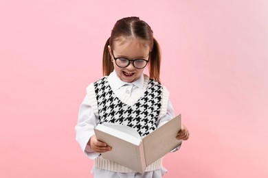 Cute little girl in glasses reading book on pink background